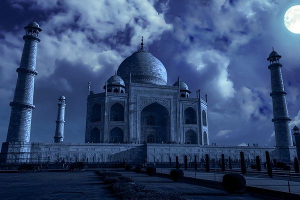 A night view of the Taj Mahal illuminated under a bright full moon, with glowing marble, dramatic clouds, and a calm, peaceful atmosphere.